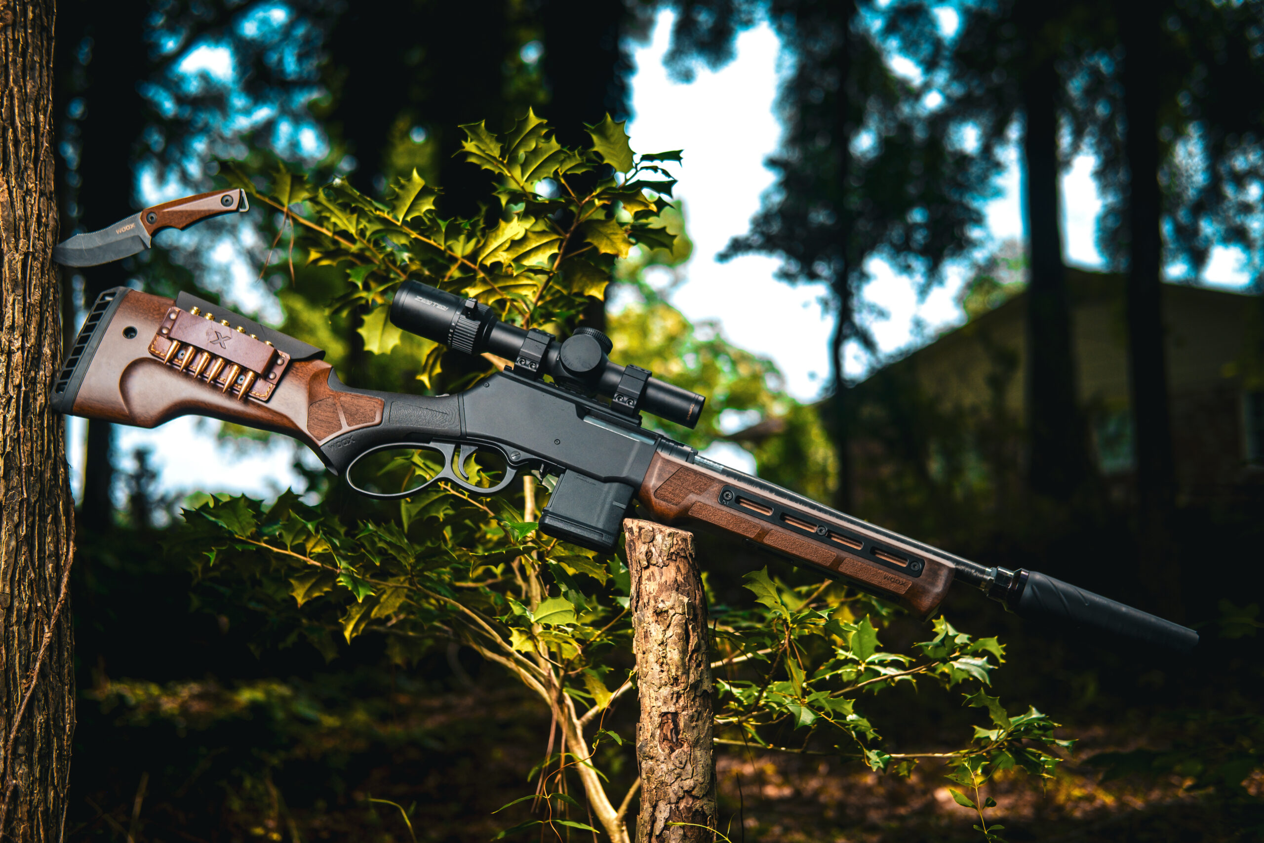 Lever-action rifle with a wooden WOOX stock, leather cartridge holder carrying six rounds, mounted riflescope, and suppressor displayed outdoors.